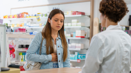 Young Woman Holding Her Stomach To Illustrate A Stomach Ache Speaking To A Pharmacist At A Pharmacy