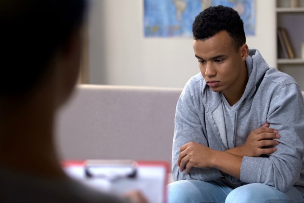 A young man looking downcast in a consulting room while being looked on by a professional holding a clipboard