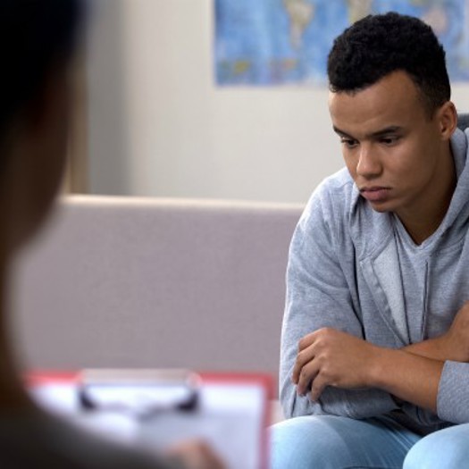A young man looking downcast in a consulting room while being looked on by a professional holding a clipboard